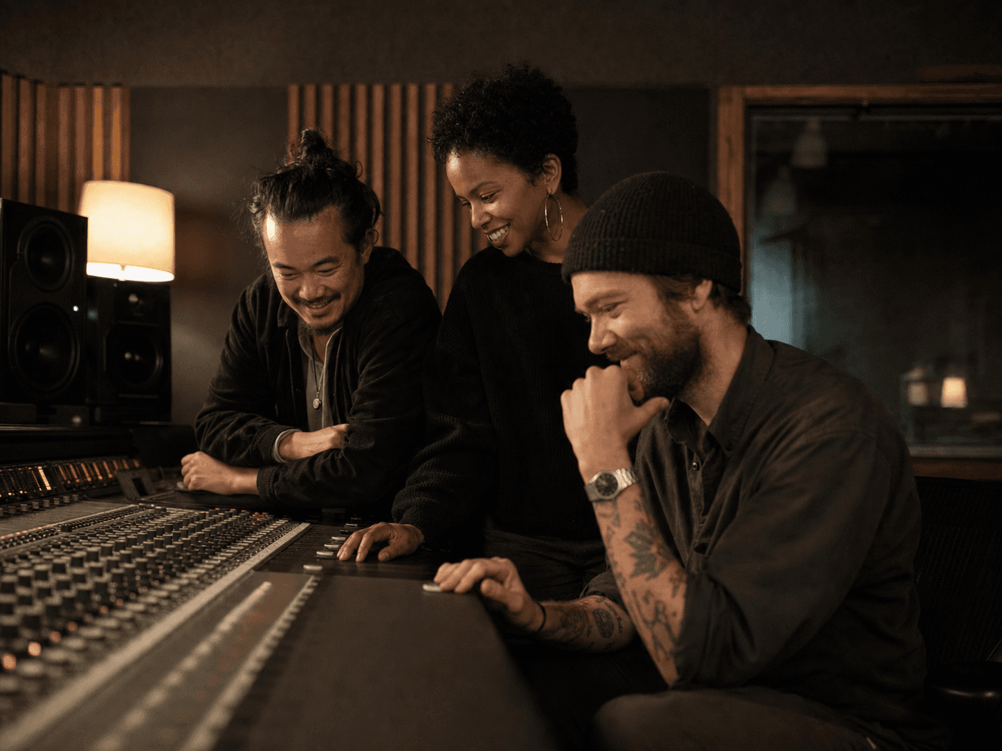Three musicians sharing a relaxed playback moment around a studio mixing console